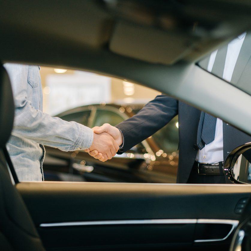 Buyer of car shaking hands with seller in auto dealership, view from interior of car. Close-up of handshake of business people. Concept of choosing and buying new car at showroom.